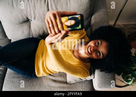 Vista dall'alto di una donna afro-americana felice che si autoritratti con il telefono cellulare mentre si sdraiano sul divano Foto Stock