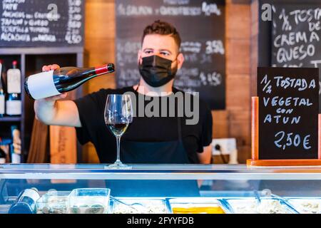 Barista anonimo versare il vino dalla bottiglia in un bicchiere sul bancone nel ristorante con molti cartelli Foto Stock
