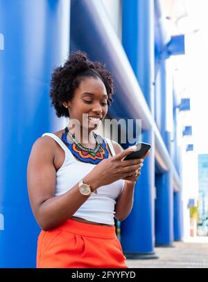 Giovane donna afro-americana allegra in estate shorts browsing mobile telefono e passeggiate in strada soleggiata Foto Stock