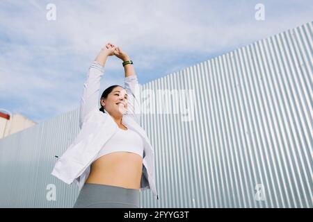 Basso angolo di felice giovane atleta etnica in abbigliamento sportivo braccia sorridenti e stretching prima di allenarsi all'aperto in città Foto Stock
