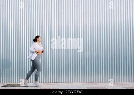 Vista laterale di un'atleta giovane e femminile di dimensioni più determinate con capelli scuri in abbigliamento sportivo che corre sulla strada della città vicino al metallo parete Foto Stock