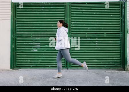 Vista laterale di un'atleta giovane e femminile di dimensioni più determinate con capelli scuri in abbigliamento sportivo che corre sulla strada della città vicino al metallo porta Foto Stock