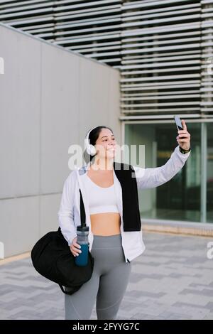 Allegra giovane donna etnica in abbigliamento sportivo e cuffie sorridenti mentre portare selfie sullo smartphone dopo l'allenamento in città Foto Stock