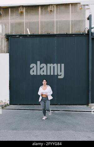 Atleta giovane e femminile con capelli scuri sportswear in esecuzione sulla strada della città vicino porta di metallo Foto Stock