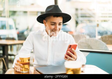 Tombino concentrato in cappello seduto al tavolo con il laptop e. bicchiere di birra mentre si scorre il telefono cellulare Foto Stock