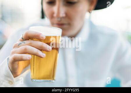 Tombino positivo corto con tatuaggi che indossa cappello e camicia bianca bere birra dal bicchiere nel caffè Foto Stock