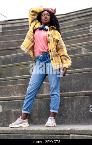 Corpo completo ad angolo basso di femmina afroamericana con wireless cuffie sul collo in piedi sulla scala in pietra con la mano testa Foto Stock