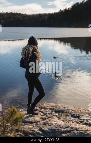 Vista posteriore corpo intero di femmina senza volto in piedi outerwear sulla costa del fiume con anatre circondato da foresta Foto Stock