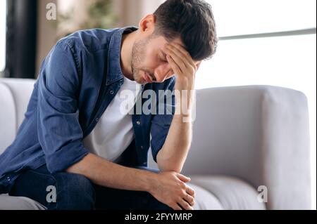Malato sconvolto stressato giovane uomo caucasico in abiti casual si siede sul divano nel soggiorno, piegandosi sopra, massaggiando la testa, sofferente di dolore, emicrania, chiudendo gli occhi, ha bisogno di cure e riposo Foto Stock