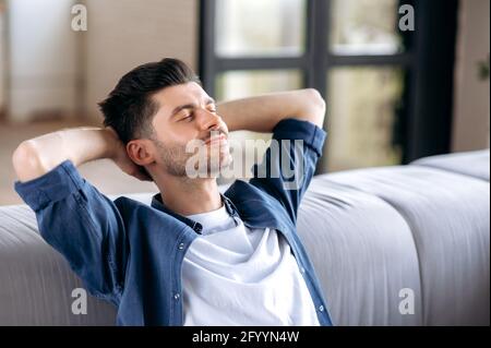 Primo piano calmo felice bell'uomo caucasico in abiti casual rilassandosi sul divano a casa, riposandosi in soggiorno, sognando di vacanza, metteva le mani dietro la testa, chiuda gli occhi, sorridendo Foto Stock