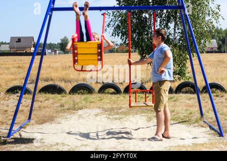 Defocalizzare la bambina che oscilla sul campo da gioco con il giovane uomo, ragazzo, suo fratello maggiore. Zona di campagna. Altalena blu e rossa. Estate in famiglia Foto Stock