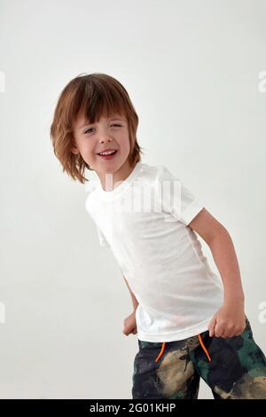 Happy boy in una t-shirt bianca casual in piedi contro il bianco parete e guardando la fotocamera Foto Stock