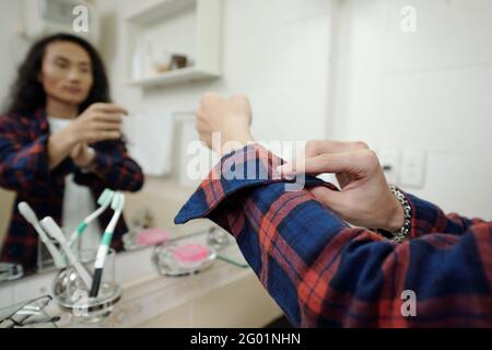 Primo piano di giovane uomo asiatico regolare camicia manica a scacchi mentre vestirsi di fronte allo specchio in bagno Foto Stock