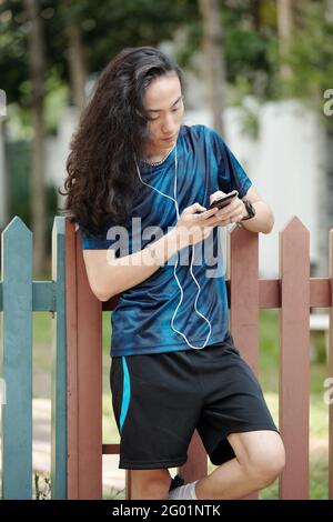 Serio giovane vietnamita dai capelli lunghi in abbigliamento sportivo in piedi al recinto e l'ascolto di audio in auricolari mentre comunica online con smartphone Foto Stock