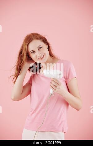 Ritratto di giovane donna sorridente con capelli rossi che le acconciano i capelli con essiccatore a soffiaggio Foto Stock