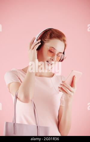 Bella giovane donna in occhiali da sole che sceglie la canzone da ascoltare nelle cuffie tramite l'applicazione sullo smartphone Foto Stock