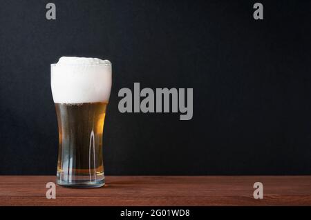 Birra fredda e schiumosa in tazza di birra di vetro su un tavolo di legno in un pub. Parete nera scura come sfondo per testo, menu, mockup Foto Stock