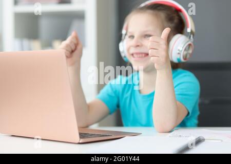 La ragazza sorridente si siede al computer portatile in cuffia e mostra i pollici su Foto Stock