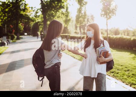 Due ragazze studentesche in maschere mediche protettive salutano i loro gomiti quando si incontrano nel campus. Formazione a distanza. Messa a fuoco selettiva morbida. Foto Stock