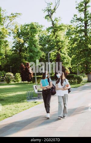 Due ragazze studentesche in maschere mediche protettive camminano e parlano nel campus. Formazione a distanza. Messa a fuoco selettiva morbida. Foto Stock