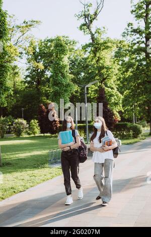 Due ragazze studentesche in maschere mediche protettive camminano e parlano nel campus. Formazione a distanza. Messa a fuoco selettiva morbida. Foto Stock