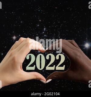 Carta di Capodanno 2022. Mani bianche e nere a forma di cuore sullo sfondo del cielo stellato e numeri 2022, cornice quadrata, spazio di copia Foto Stock
