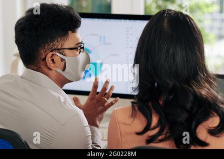 Giovane uomo d'affari in maschera protettiva che mostra il diagramma circolare sul computer schermo al collega e spiegare i suoi dettagli importanti Foto Stock