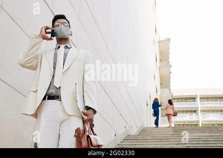 Bel giovane uomo d'affari in vestito bianco e maschera medica che scende le scale lasciando l'edificio dell'ufficio e parlando al telefono con un collega o un partner commerciale Foto Stock