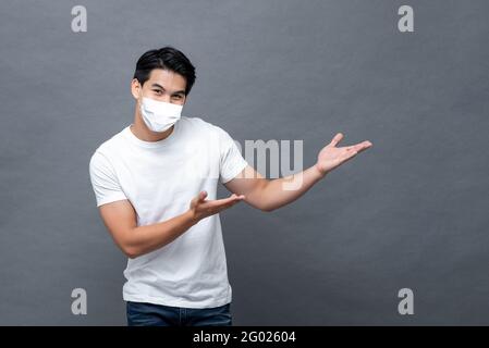 Giovane uomo asiatico che indossa maschera facciale aprendo le mani a vuoto spazio da parte in isolato sfondo grigio chiaro studio Foto Stock