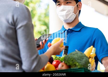 Uomo di deliveryman asiatico igienico che indossa la maschera facciale mentre consegna i generi alimentari al cliente a casa, consegna di cibo nel momento del concetto pandemico Foto Stock