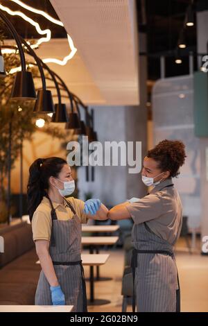 Ritratto verticale di due giovani donne che saltano i gomiti come un saluto senza contatto mentre si lavora in un bar, concetto di sicurezza covid Foto Stock