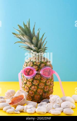 Ananas in occhiale da nuoto rosa con ciottoli di mare e conchiglie primo piano su sfondo giallo-blu, concetto estivo Foto Stock