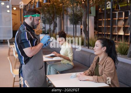 Vista laterale ritratto del cameriere che indossa visiera e guanti mentre si prende ordine in caffetteria con le misure di sicurezza covid, copy space Foto Stock