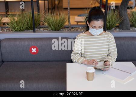 Ritratto della giovane donna asiatica che indossa la maschera e usa lo smartphone mentre si lavora al tavolo nel caffè Foto Stock
