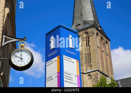 Viersen (Suchteln), Germania - Maggio 20. 2021: Vista sulla colonna di informazioni turistiche con orologio e la torre della chiesa sfondo nel villaggio tedesco (fuoco su pillola Foto Stock