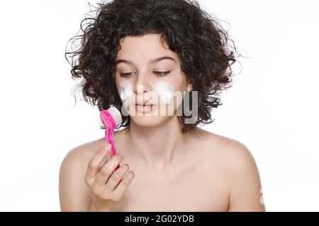 Skincare adolescente. Bella ragazza teenage con i capelli ricci gorgeous usando spazzola del viso e pulitore schiumogeno. Concetto di lavaggio del viso isolato su sfondo bianco Foto Stock