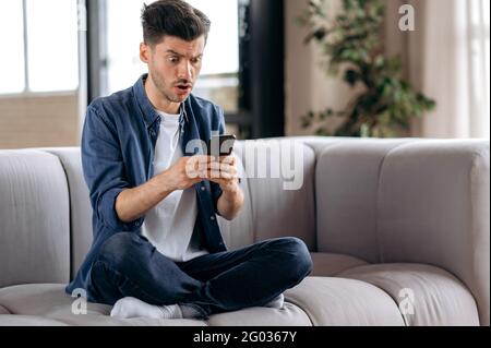 Scioccato confuso giovane uomo caucasico, in abbigliamento casual, seduto sul divano in posizione lotus, utilizza smartphone, ricevendo un messaggio inatteso, testando con gli amici, guardando lo schermo a sorpresa Foto Stock