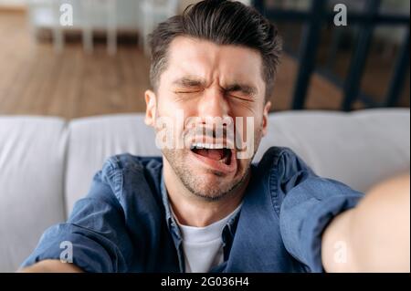 Selfie di un triste turbato giovane caucasico con una barba, in abiti casual, grimacing e imbrogliare in giro mentre prendendo selfie, usa uno smartphone, volto ritorto, occhi chiusi Foto Stock