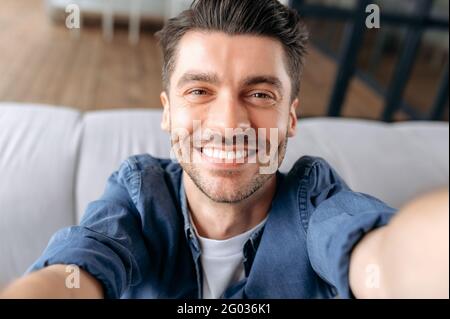 Giovane adulto caucasico uomo prende un selfie. Ritratto di simpatico ragazzo allegro, in camicia denim, seduto sul divano, tenendo lo smartphone in mano e guardando la webcam, sorridente amichevole Foto Stock