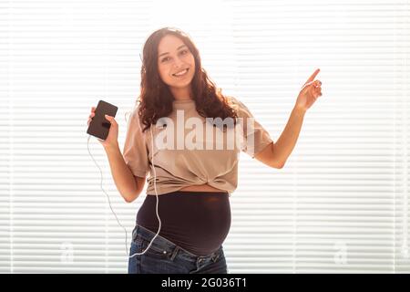 Una donna incinta pacificata con capelli ricci ascolta la piacevole musica classica con smartphone e cuffie. Concetto di un umore rilassante prima Foto Stock