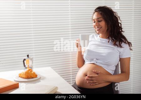 Gioiosa giovane incinta bella donna guarda attraverso le persiane durante la sua colazione mattutina con caffè e croissant. Concetto di Buongiorno e. Foto Stock