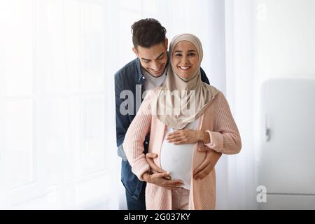 Amorevole marito arabo abbracciando la sua moglie musulmana incinta mentre si è in piedi vicino alla finestra Foto Stock