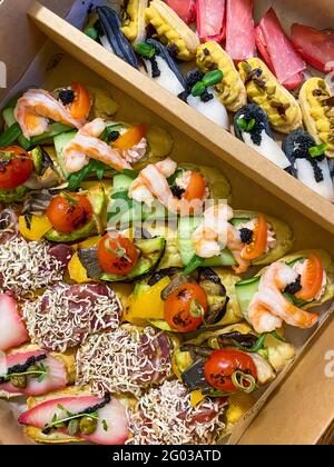 Scatola con piccoli panini, eclair, bruschetta con affettati, verdure alla griglia, formaggio e frutti di mare. Tavole di salumi da ordinare. Foto Stock