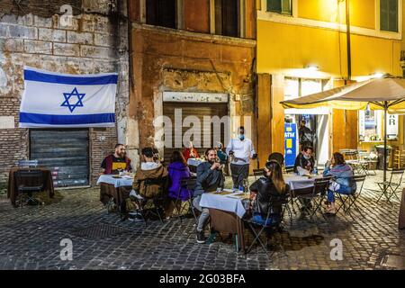 La gente cena fuori di un ristorante nel ghetto ebraico in Roma di notte con una bandiera israeliana appesa al muro Foto Stock