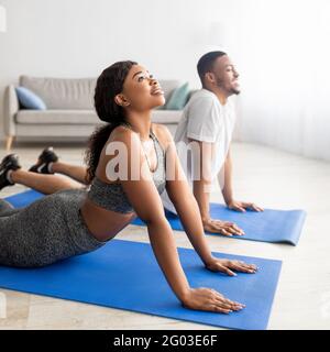 Concetto sportivo domestico. Flessibile coppia nera in abbigliamento sportivo che fa yoga cobra asana sul tappetino a casa Foto Stock