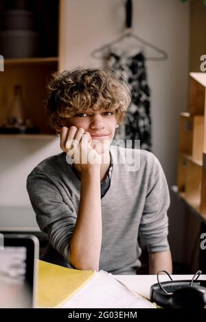 Ritratto di ragazzo sorridente seduto con la mano sul mento alla scrivania Foto Stock