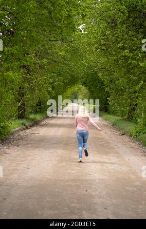 La bionda ragazza sulla strada corre in un arco di albero, verde Foto Stock