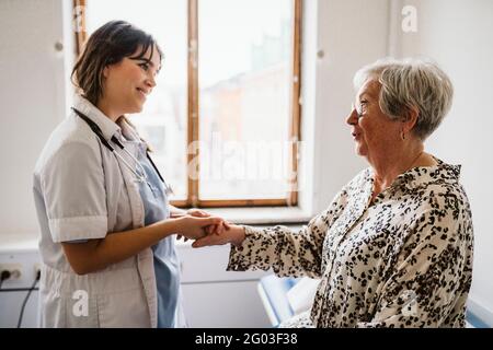 Sorridente operatrice sanitaria che guarda pazienti anziani parlare mentre si tiene le mani Foto Stock