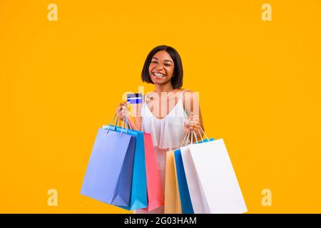 Shopping stagionale. Donna nera sorridente che tiene borse regalo e carta di credito, sorridendo alla macchina fotografica su sfondo arancione studio Foto Stock