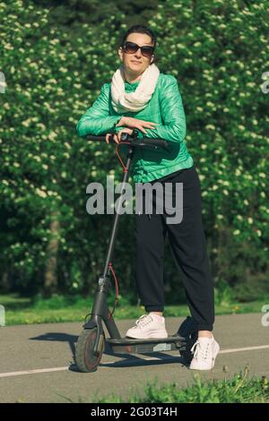 La giovane donna si fissa con le mani piegate sul volante di uno scooter elettrico nel parco verde Foto Stock